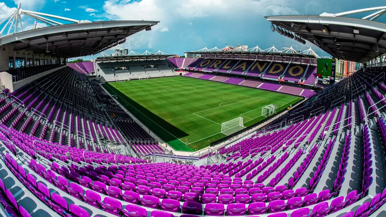 Empty soccer stadium with green field and purple seats. The word "Orlando" is visible on the upper seats. Overhead roof structure covers the seating areas. Bright, partly cloudy sky above.