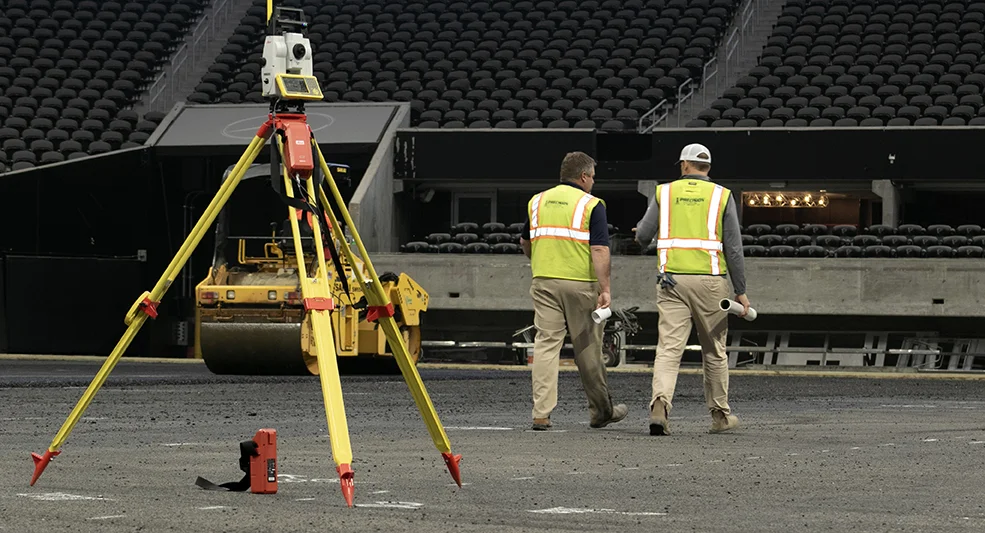 Two construction workers from a sports turf company, wearing safety vests and helmets, walk across a gravel surface in a stadium. Surveying equipment is set up in the foreground.